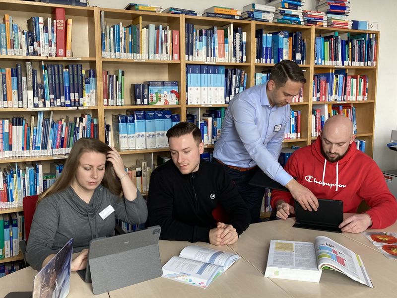 Gruppe von Schüler*innen sitzt in Bibliothek der Pflegeschule, mit Büchern und Tablet