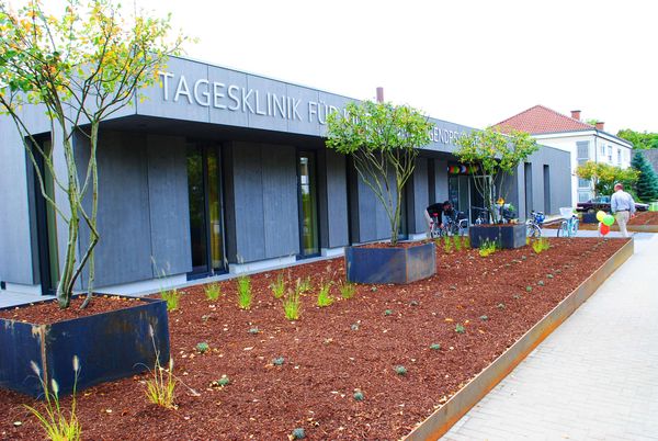 Blick auf die grau-blaue Außenfassade der Tagesklinik, davor ein Beet mit Pflanzen und Bäumen