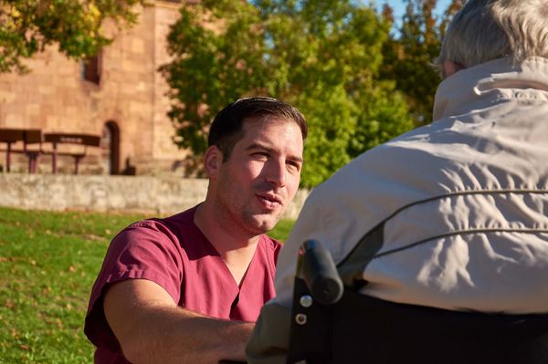In einem Garten kniet bzw. hockt ein Mitarbeiter vor einem Patienten im Rollstuhl und spricht mit ihm
