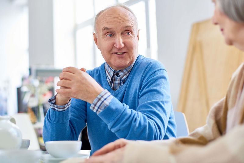 Ein älterer Herr in einem blauen Pullover sitzt mit einer älteren Dame am Tisch und unterhält sich