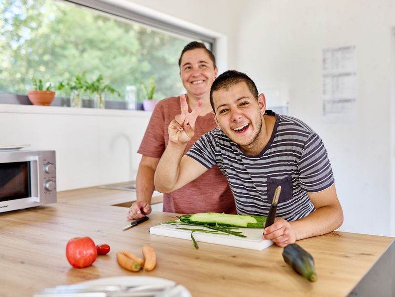 Klient kocht mit Assistenzkraft, zeigt lachend Victory-Zeichen mit der Hand