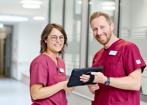 Zwei Pflegekräfte stehen mit Tablet auf einem Stationsflur und lächeln in die Kamera