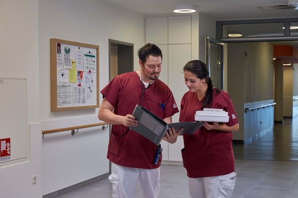 Zwei Pflegefachkräfte in Arbeitskleidung blicken im Krankenhausflur gemeinsam auf Unterlagen